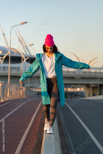 Stylish happy young woman wearing sports leggings, white sneakers, bright hat smiling enjoy her weekends, walking on curb near road. Charming hipster dressed in casual outfit outdoor at sunny light. 