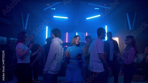 A pretty girl with a cocktail and a phone in her hands is dancing, a crowd of people are dancing around the girl on the dance floor of a nightclub under the light of colored spotlights. Night club