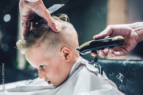 Lifestyle photo. Barber shop little blond boy customer. Hairdresser man work move hand comb tool scissors trimmer white cape. Modern style kid short hair cut. Fashion business service for child