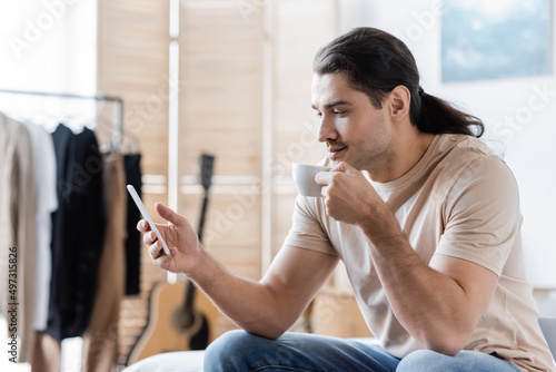 man with long hair holding cup of coffee and using mobile phone.