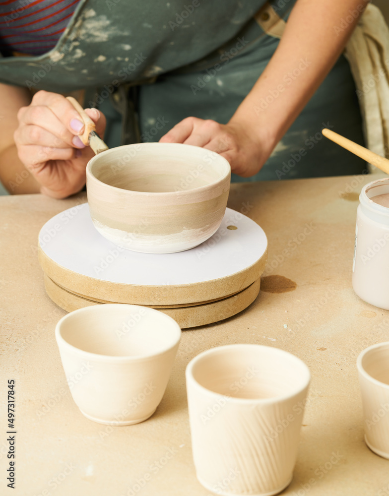 Close-up of girl painting clay mug with glaze. Woman coloring pottery in workshop with a paintbrush. Painter in green apron glazing clay pot. Close-up of girl painting clay mug with glaze. Woman coloring pottery in workshop with a paintbrush. Painter in green apron glazing clay pot.