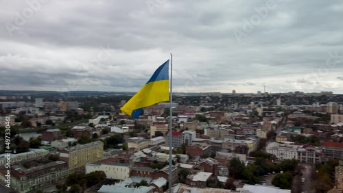 Flagpole with flag of Ukraine with epic gray sky. Counterclockwise city aerial view above river Lopan near Skver Strilka and Holy Annunciation Cathedral in Kharkiv downtown, Ukraine photo