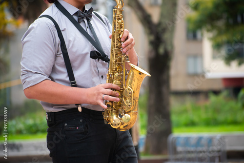 Man in blues or jazz suit, with black tie and black pants playing a saxophone in public in a public park. 