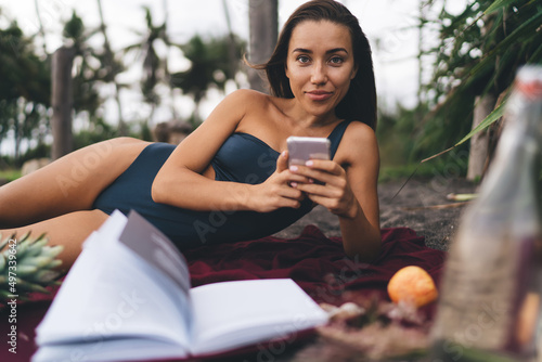 Portrait of millennial female in swimsuit using cellular device while tan sunbathing at seashore beach, young Caucaisan travel blogger with mobile gadget looking at camera lying at coastline #497339642