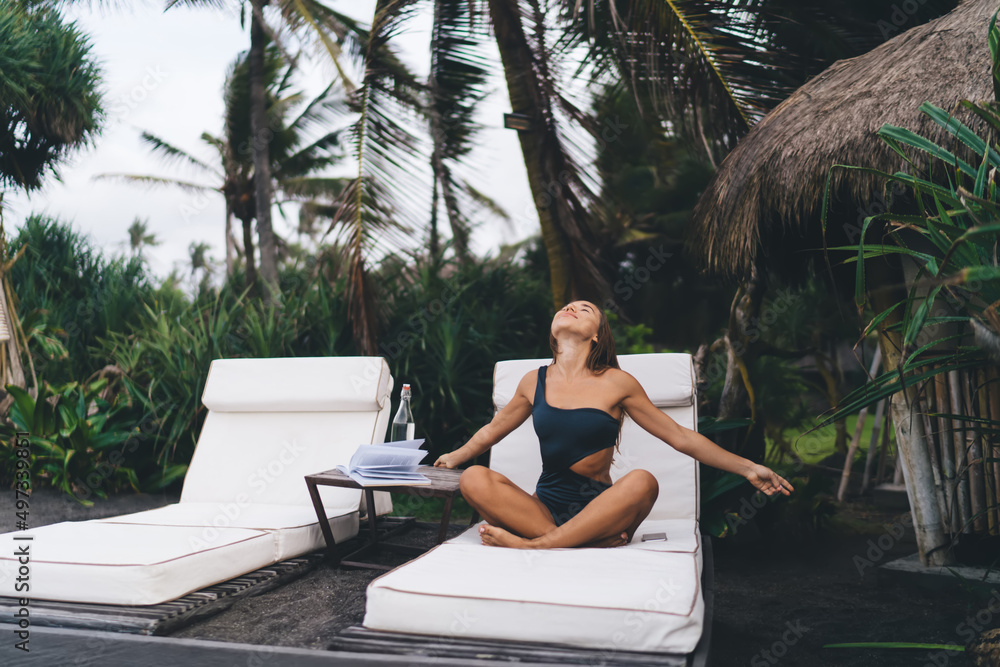 Carefree hipster girl in swimwear recreating at comfortable sunbed enjoying travel vacations at tropical island, woman with good figure and literature raising hands in lounge zone with closed eyes