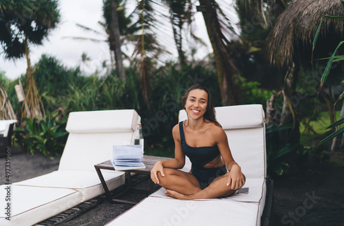 Cheerful female reader in swimsuit with literature best seller smiling during leisure pastime in lounge zone of coastline beach, happy woman with novel poetry book enjoying hobby time at seashore