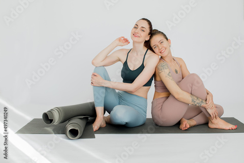 The girls smile and rest after yoga classes on the mat leaning on each other. Sunbeams illuminate the white studio