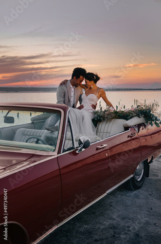 Beautiful couple at sunset on a summer wedding day. Romantic couple in love next to their convertible. The groom is in a light suit, the bride is in a beautiful stylish dress. © ann