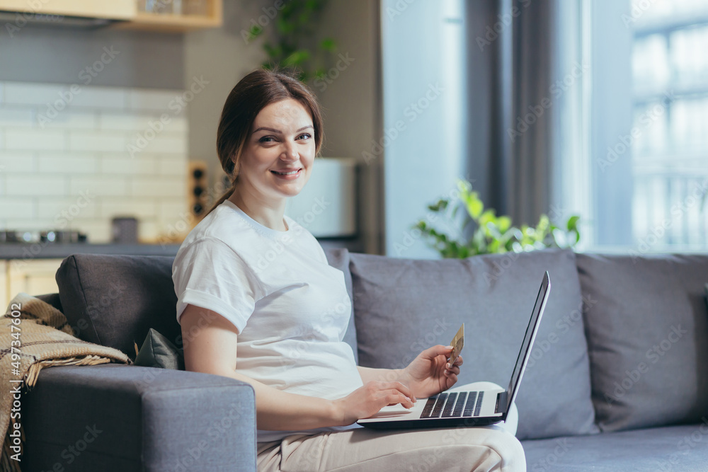 Fototapeta premium Pregnant woman sitting on sofa at home, using laptop for shopping in online store, looking at camera and smiling holding bank credit card