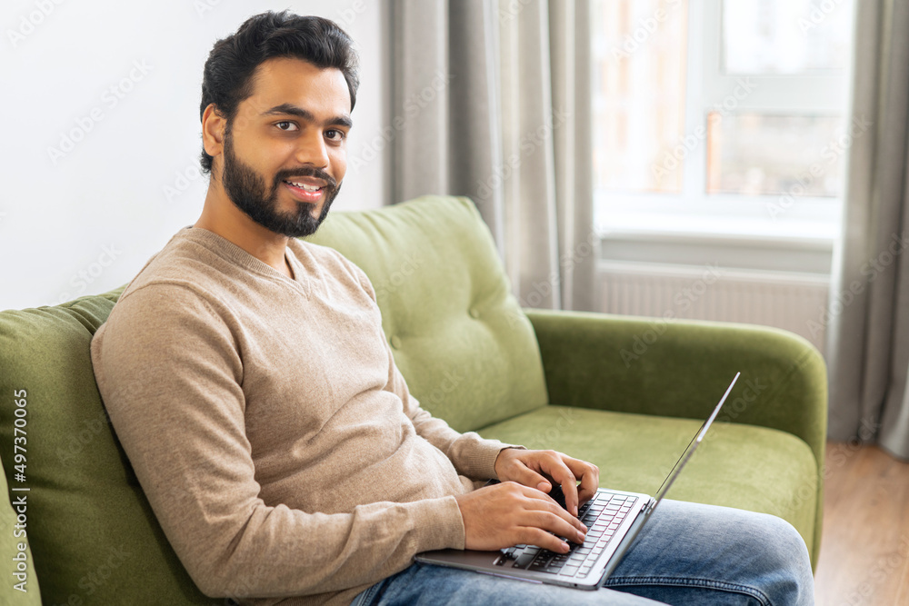 Young Indian male student watching webinars, educational courses, learning on the distance. Front view of positive Hindi man in smart casual clothes using laptop while sitting at the sofa in his flat Young Indian male student watching webinars, educational courses, learning on the distance. Front view of positive Hindi man in smart casual clothes using laptop while sitting at the sofa in his flat