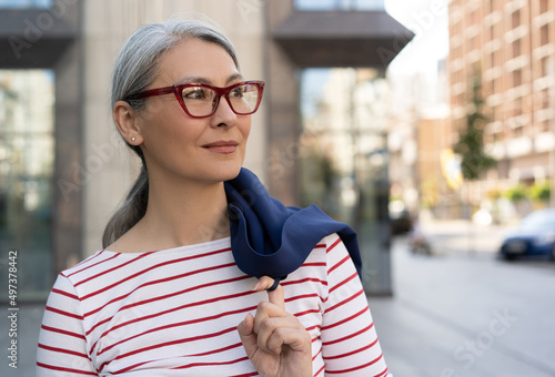 Confident mature businesswoman wearing stylish eyeglasses standing on urban street, copy space. Portrait of pensive asian female looking away waiting for taxi outdoors