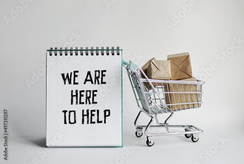 A notebook with the inscription WE ARE HERE TO HELP. With a decorative grocery cart along with small packaged mailboxes in the background. 