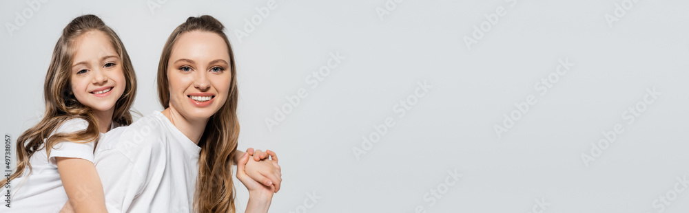 pleased mother and daughter in white t-shirts holding hands and smiling at camera isolated on grey, banner. pleased mother and daughter in white t-shirts holding hands and smiling at camera isolated on grey, banner.