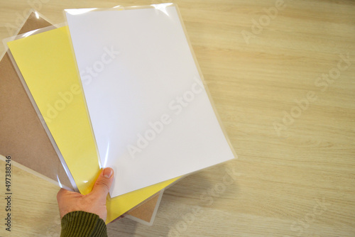 Worker grabbing some laminating pouches with yellow, craft and white sheets inside the plastic, and blank space on the right.