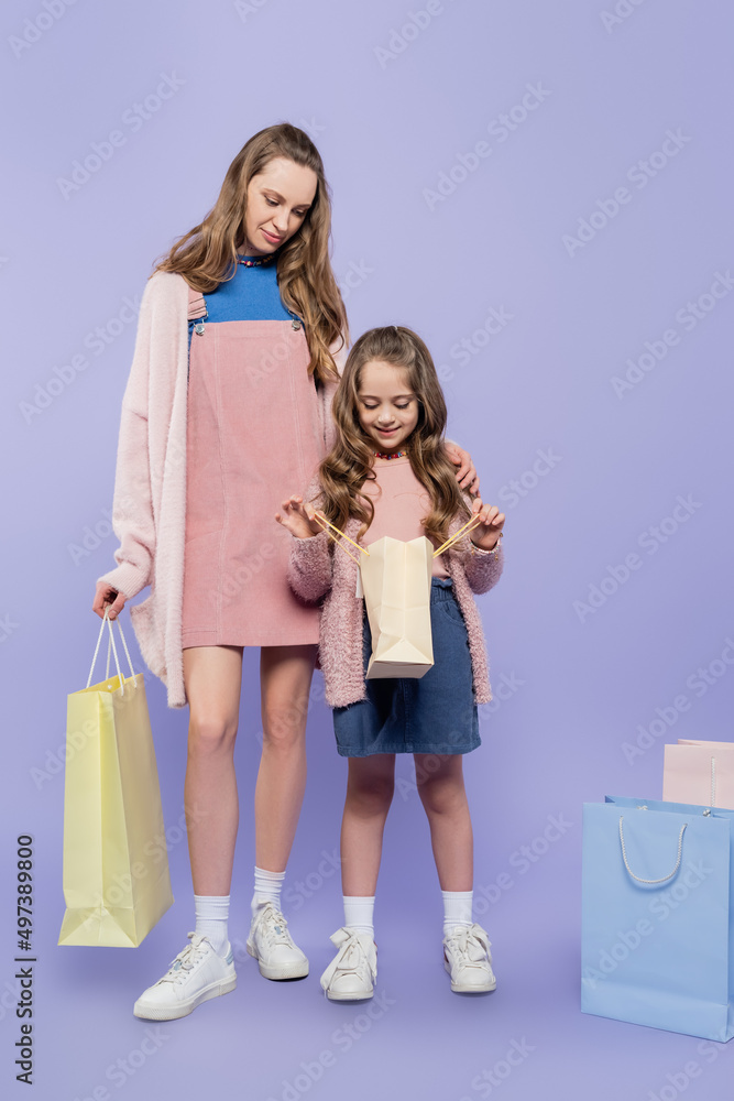 full length of kid looking inside of shopping bag near mother on purple. full length of kid looking inside of shopping bag near mother on purple.