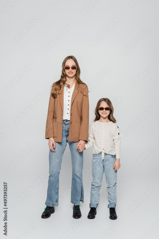 full length view of trendy mother and daughter in sunglasses holding hands on grey background. full length view of trendy mother and daughter in sunglasses holding hands on grey background.