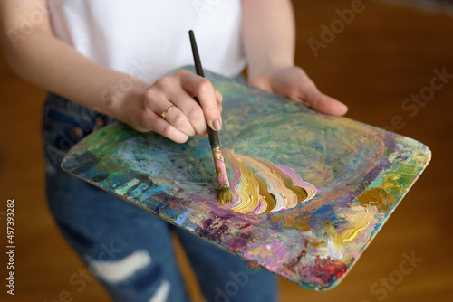 Wooden palette close-up. The artist girl mixes various shades of bright colored oil or gouache paints. Wavy strokes of paint with a brush on a wooden board. Artist's home studio. Close-up, soft focus.