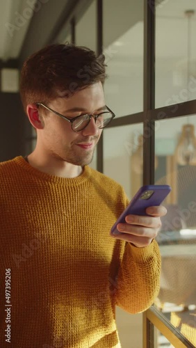 A handsome young student stands near the window, keeps a phone in his hands and reads something thoughtfully
