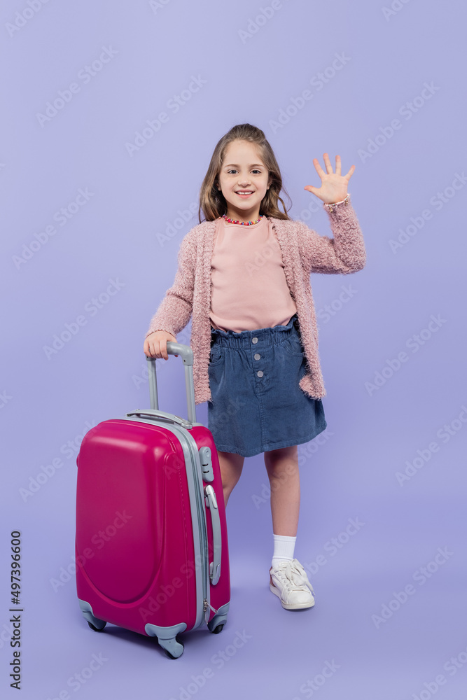 full length of happy girl waving hand near pink baggage on purple. full length of happy girl waving hand near pink baggage on purple.