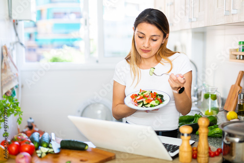 Young peruvian housewoman eating vegetable salad and watching movie on laptop