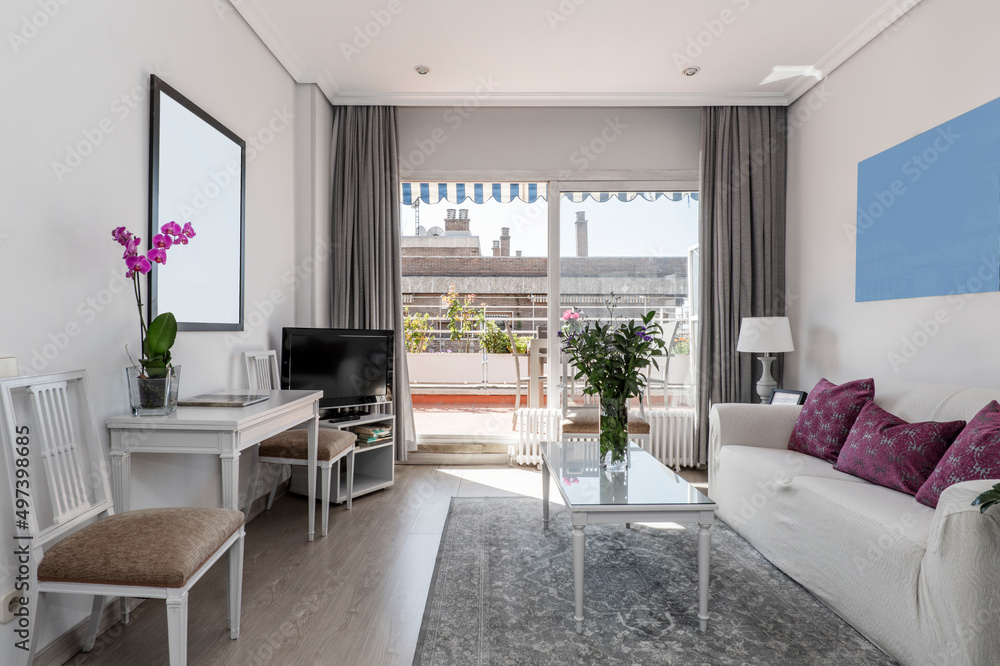 living room with three-seater sofa, white wooden furniture, tv on a table, gray carpet and access to a terrace with plants and flowers living room with three-seater sofa, white wooden furniture, tv on a table, gray carpet and access to a terrace with plants and flowers