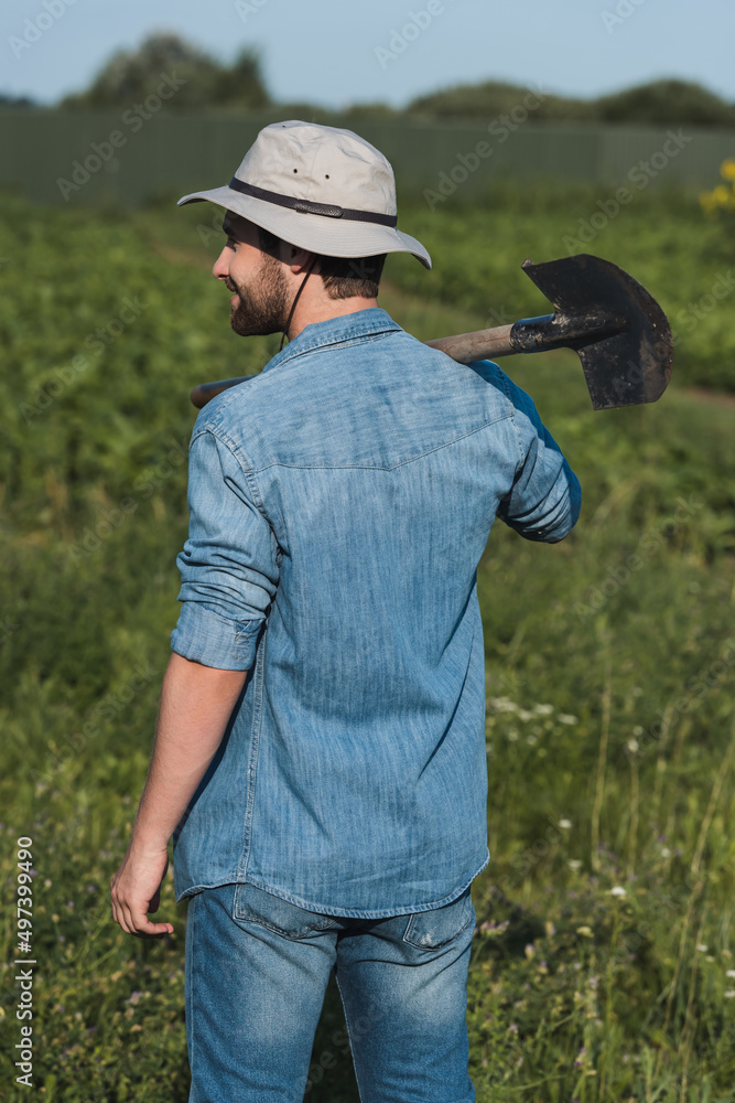 back view of farmer in denim shirt and brim hat standing with shovel in field. back view of farmer in denim shirt and brim hat standing with shovel in field.