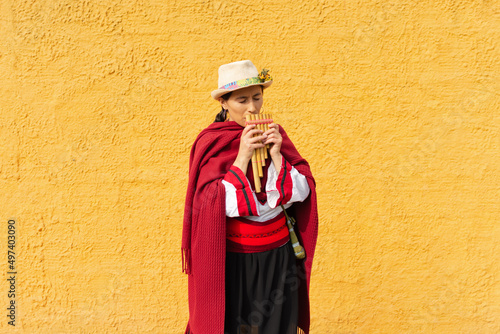 Portrait of an indigenous woman with a hat and typical costume playing an Andean panpipe with a rustic yellow wall in the background photo