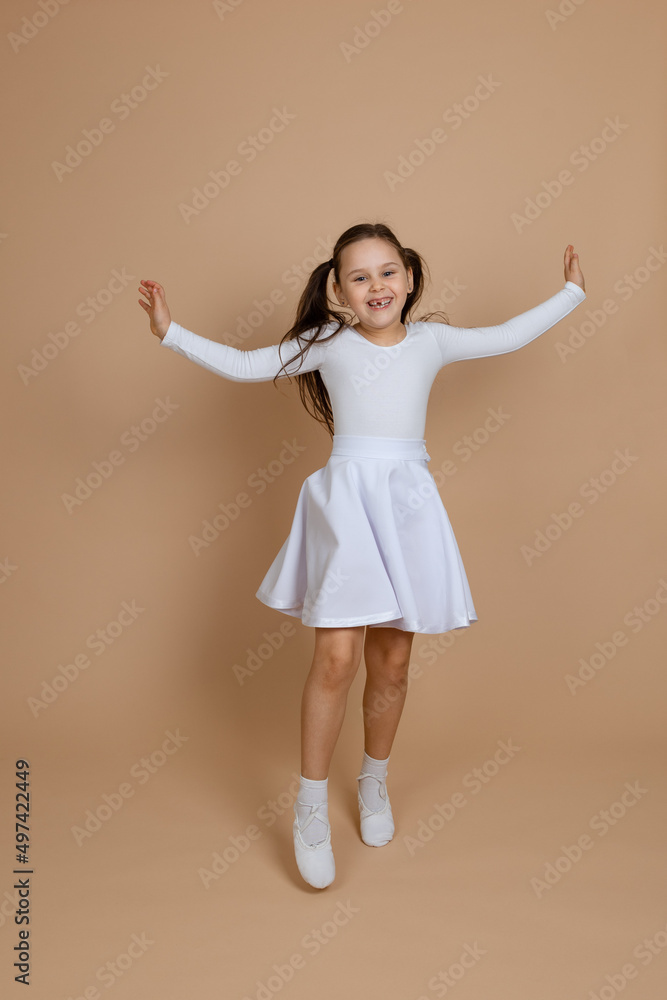 Young cute smiling girl with long dark hair in white dress, socks and gymnastics shoes dancing, spreading hands, raising leg, having fun on brown background. Ballet, gymnastics, sport, cheerleading. Young cute smiling girl with long dark hair in white dress, socks and gymnastics shoes dancing, spreading hands, raising leg, having fun on brown background. Ballet, gymnastics, sport, cheerleading.