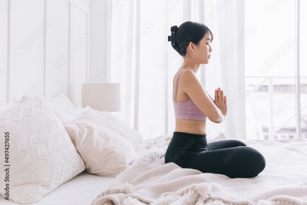 Young smiling attractive sporty Asian woman practicing yoga on the bed, doing Ardha Padmasana exercise, meditating in Half Lotus pose with namaste, indoor working out at home, wearing sportswear. Young smiling attractive sporty Asian woman practicing yoga on the bed, doing Ardha Padmasana exercise, meditating in Half Lotus pose with namaste, indoor working out at home, wearing sportswear.