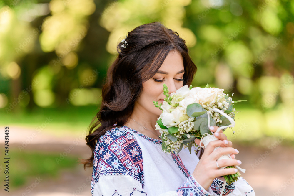 Close view of beautiful and cute bride with long curly hair and natural makeup, wearing in embroidery dress with colorful pattern, holding wedding bouquet and smelling it during posing on nature Close view of beautiful and cute bride with long curly hair and natural makeup, wearing in embroidery dress with colorful pattern, holding wedding bouquet and smelling it during posing on nature