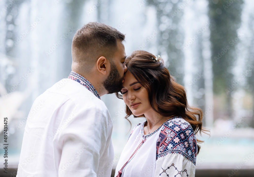Close view of stunning and pensive woman with natural makeup and curly hair, dressed in shirt with colorful pattern, bending head forward while man kissing her face on background of fountains Close view of stunning and pensive woman with natural makeup and curly hair, dressed in shirt with colorful pattern, bending head forward while man kissing her face on background of fountains