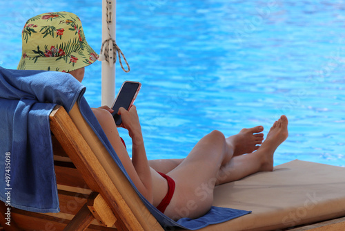 Slim girl in bikini lying with smartphone in hands in deck chair near swimming pool in shadow of sun umbrella. Summer holiday and online communication at resort © Oleg