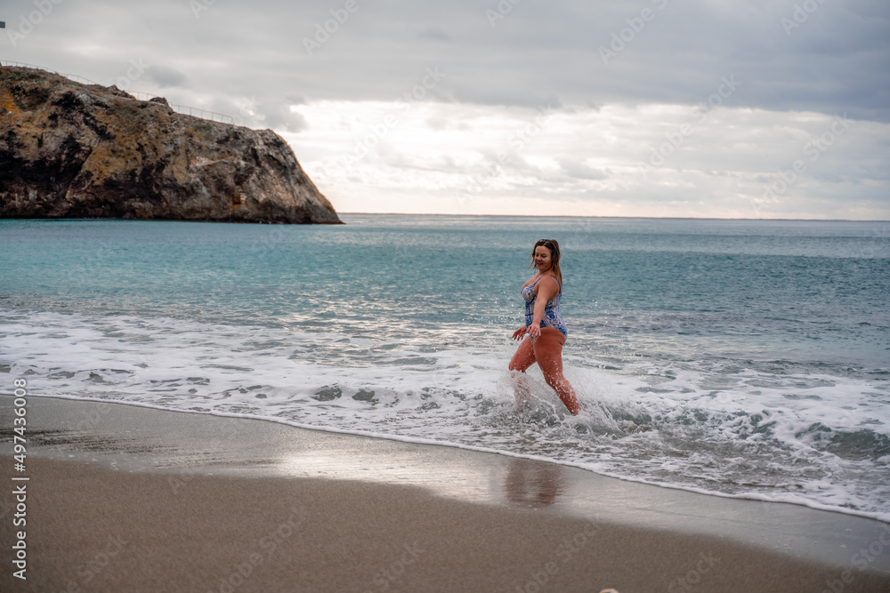 A plump woman in a bathing suit enters the water during the surf. Alone on the beach, Gray sky in the clouds, swimming in winter. A plump woman in a bathing suit enters the water during the surf. Alone on the beach, Gray sky in the clouds, swimming in winter.