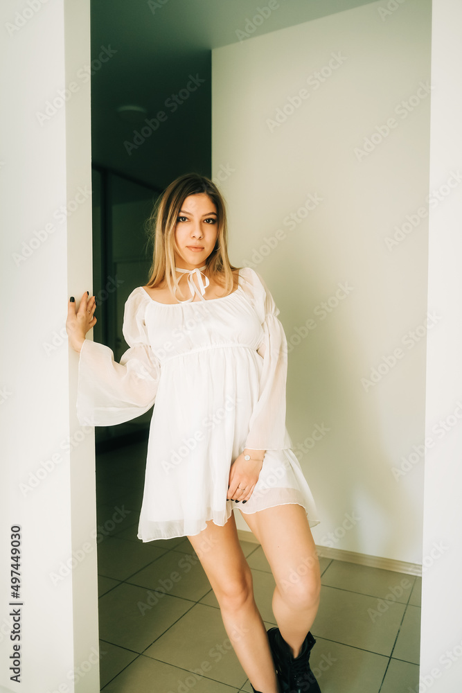 Portrait of attractive caucasian young woman in a white dress standing in a empty room with white walls.