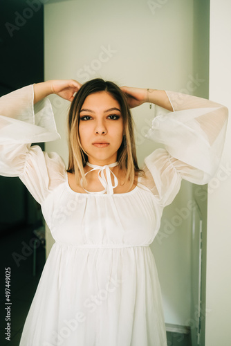 Portrait of attractive caucasian young woman in a white dress standing in a empty room with white walls.