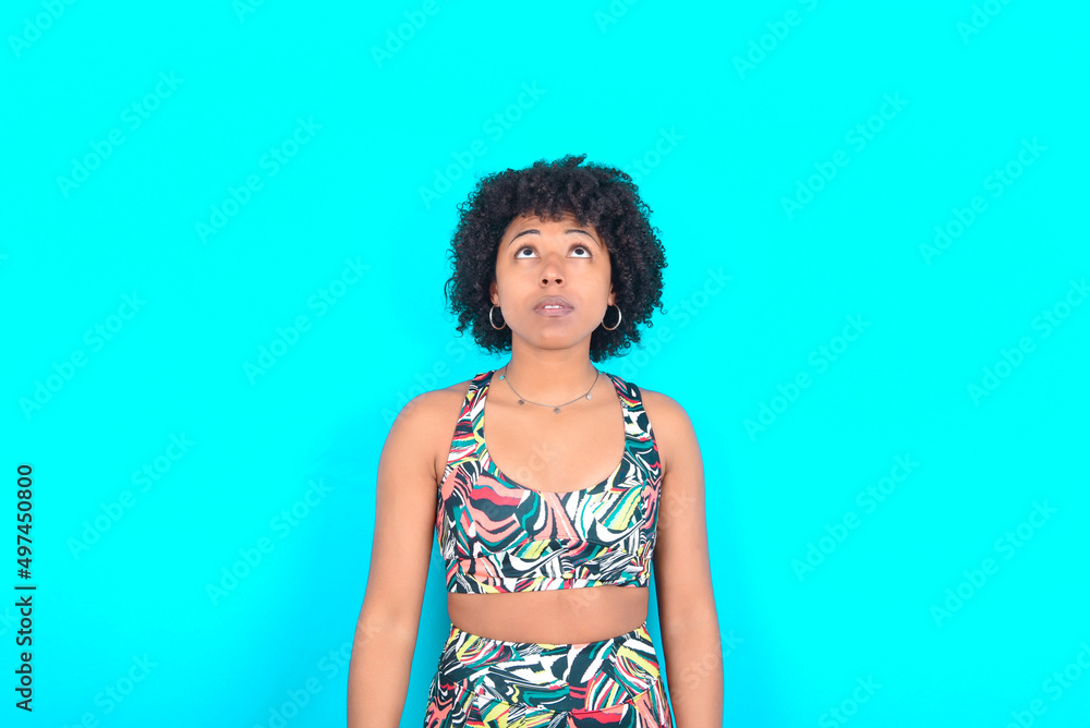 young woman with afro hairstyle in sportswear against blue background looking up as he sees something strange. young woman with afro hairstyle in sportswear against blue background looking up as he sees something strange.