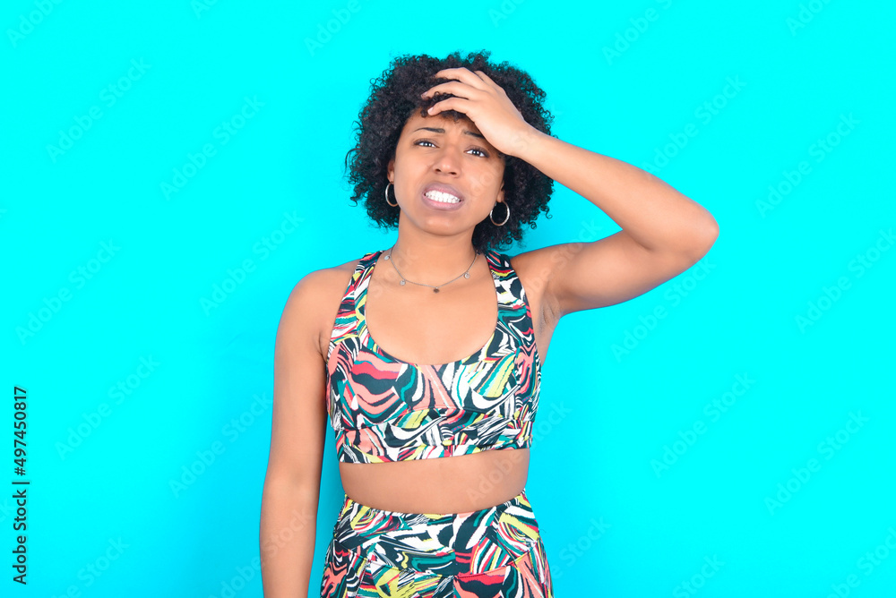 Oops, what did I do? young woman with afro hairstyle in sportswear against blue background holding hand on forehead with frightened and regret expression. Oops, what did I do? young woman with afro hairstyle in sportswear against blue background holding hand on forehead with frightened and regret expression.