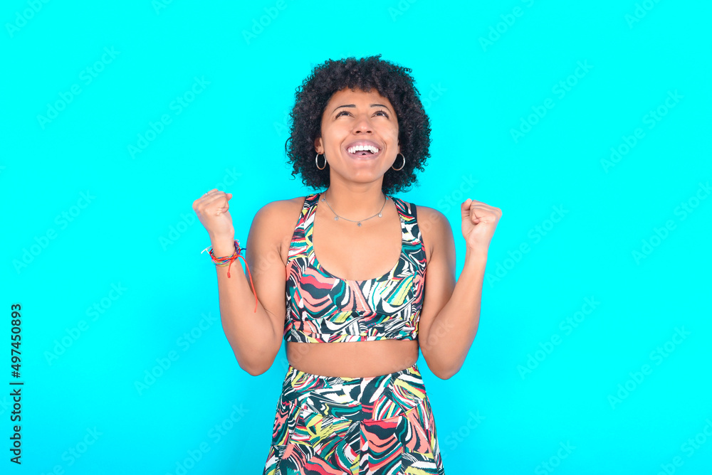 young woman with afro hairstyle in sportswear against blue background looks with excitement up, keeps hands raised, notices something unexpected. young woman with afro hairstyle in sportswear against blue background looks with excitement up, keeps hands raised, notices something unexpected.