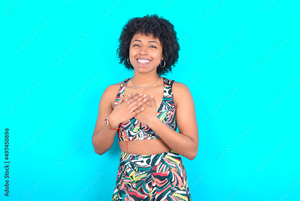 Honest young woman with afro hairstyle in sportswear against blue background keeps hands on chest, touched by compliment or makes promise, looks at camera with great pleasure. Honest young woman with afro hairstyle in sportswear against blue background keeps hands on chest, touched by compliment or makes promise, looks at camera with great pleasure.