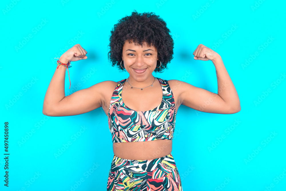 Waist up shot of young woman with afro hairstyle in sportswear raises arms to show muscles feels confident in victory, looks strong and independent, smiles positively at camera. Sport concept. Waist up shot of young woman with afro hairstyle in sportswear raises arms to show muscles feels confident in victory, looks strong and independent, smiles positively at camera. Sport concept.