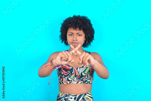young woman with afro hairstyle in sportswear against blue background has rejection angry expression crossing fingers doing negative sign.