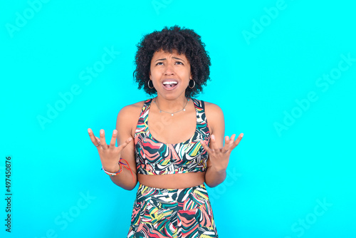 young woman with afro hairstyle in sportswear against blue background crying and screaming. Human emotions, facial expression concept. Screaming, hate, rage.