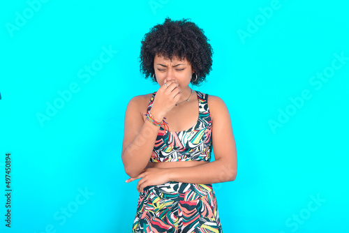 young woman with afro hairstyle in sportswear against blue background , holding his nose because of a bad smell.