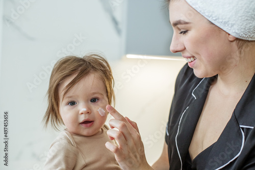 Mom puts baby cream on face of toddler daughter. Cozy house. Quality baby cosmetics.