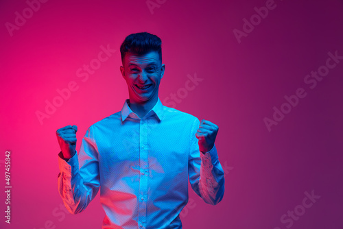 Excited young stylish man, student isolated over magenta color studio background in blue neon light. Concept of emotions, menatl health, news photo