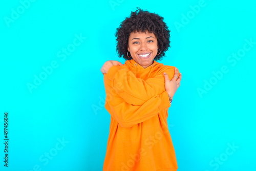 Charming pleased young woman with afro hairstyle wearing orange hoodie against blue background embraces own body, pleasantly feels comfortable poses. Tenderness and self esteem concept © Jihan