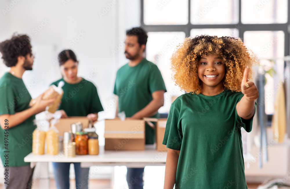 Obraz premium charity, donation and volunteering concept - happy smiling female volunteer pointing to camera and international group of people packing food in boxes at distribution or refugee assistance center