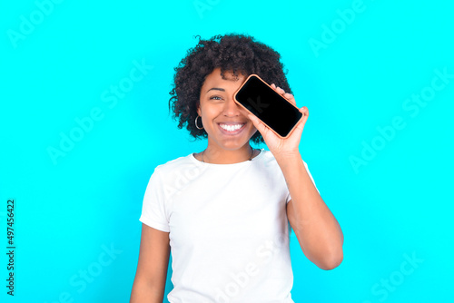 young woman with afro hairstyle wearing white T-shirt against blue wall holding modern smartphone covering one eye while smiling