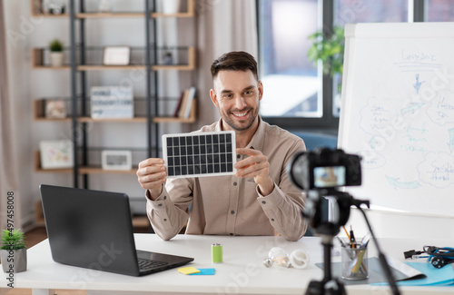 distance education, school and green energy concept - happy smiling male teacher with camera and solar battery model having online class at home office