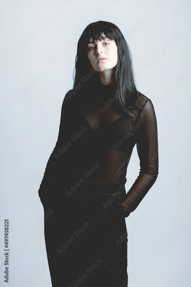 Fashion and beauty concept. Studio portrait of beautiful woman with black long hair and outfit standing and looking to camera with her hands in pockets. Grey background. Soft blue filter applied Fashion and beauty concept. Studio portrait of beautiful woman with black long hair and outfit standing and looking to camera with her hands in pockets. Grey background. Soft blue filter applied